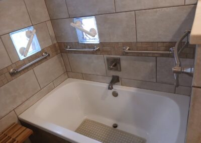 Bathtub area with multiple stainless Grab bars along tiled walls, a central faucet, and a textured non-slip mat inside the tub, plus a small wooden bench nearby.