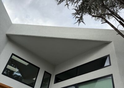 Modern white building corner with large black-framed windows and a wooden door beneath an overhang, against a cloudy sky and tree above.